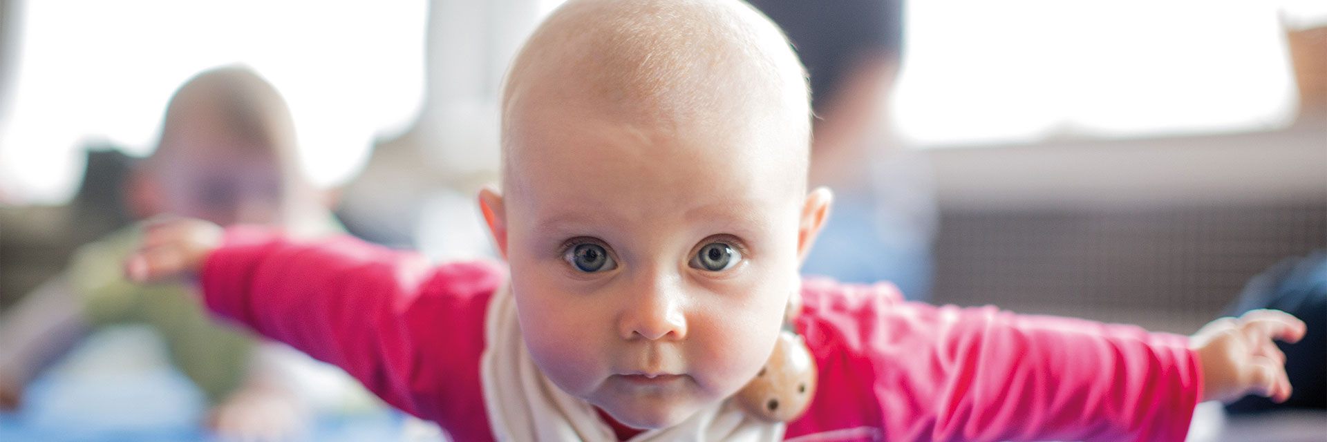 Foto: Ein Baby liegt auf dem Bauch und blickt in die Kamera. Es hat seine Arme weit zur Seite ausgestreckt.