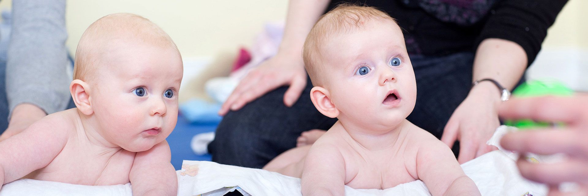 Foto: Zwei Babys liegen auf dem Bauch auf einer Wickelunterlage. Sie schauen das grüne Spielzeug in der Hand eines Erwachsenen mit großen Augen an.