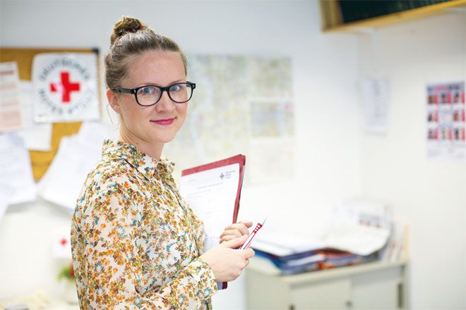 Foto: Eine junge Mitarbeiterin des DRK-Suchdienstes blickt freundlich und aufgeschlossen in die Kamera. Sie hält einen Stift sowie eine Mappe mit Formularen in den Händen.